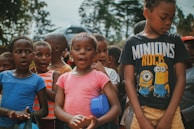A group of children is gathered outdoors, engaged with an unseen activity. The kids are wearing casual clothes in various colors, including blue, pink, orange, and black. One child is wearing a 'Minions Rock' shirt. The background features trees and there is a blurred car in the distance, suggesting a park or outdoor setting.