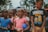 A group of children is gathered outdoors, engaged with an unseen activity. The kids are wearing casual clothes in various colors, including blue, pink, orange, and black. One child is wearing a 'Minions Rock' shirt. The background features trees and there is a blurred car in the distance, suggesting a park or outdoor setting.