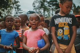A group of children is gathered outdoors, engaged with an unseen activity. The kids are wearing casual clothes in various colors, including blue, pink, orange, and black. One child is wearing a 'Minions Rock' shirt. The background features trees and there is a blurred car in the distance, suggesting a park or outdoor setting.