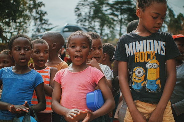 A group of children is gathered outdoors, engaged with an unseen activity. The kids are wearing casual clothes in various colors, including blue, pink, orange, and black. One child is wearing a 'Minions Rock' shirt. The background features trees and there is a blurred car in the distance, suggesting a park or outdoor setting.