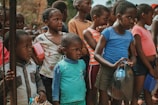 A group of children are gathered closely together, some holding containers and bags. They appear to be standing in a line or waiting for something. The children are dressed in casual clothing with a variety of colors and patterns.