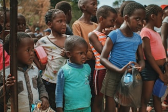 A group of children are gathered closely together, some holding containers and bags. They appear to be standing in a line or waiting for something. The children are dressed in casual clothing with a variety of colors and patterns.