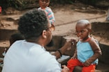 A caregiver is feeding a young child with a spoon while sitting outdoors. The child is wearing a blue and brown shirt with red pants and looks contemplative. Another child is nearby wearing a striped shirt, observing the scene. The setting appears to be an informal outdoor space with dirt and concrete steps.