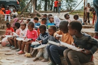 A group of young children sit on stone steps, holding white food trays as they eat. They are outdoors, with trees and a fence in the background. The children wear colorful clothing and appear engaged in their meals or in conversation with one another. A few other children and adults are visible in the background, standing near a parked car.