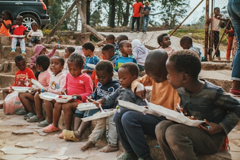 A group of young children sit on stone steps, holding white food trays as they eat. They are outdoors, with trees and a fence in the background. The children wear colorful clothing and appear engaged in their meals or in conversation with one another. A few other children and adults are visible in the background, standing near a parked car.