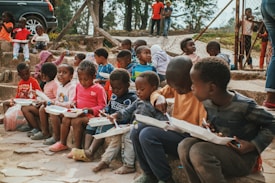 A group of young children sit on stone steps, holding white food trays as they eat. They are outdoors, with trees and a fence in the background. The children wear colorful clothing and appear engaged in their meals or in conversation with one another. A few other children and adults are visible in the background, standing near a parked car.