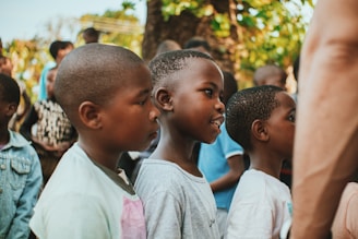 A group of children standing in a line, outdoors, with lush greenery in the background. The children appear to be engaged or focused, possibly watching something or someone.