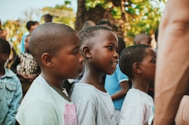 A group of children standing in a line, outdoors, with lush greenery in the background. The children appear to be engaged or focused, possibly watching something or someone.