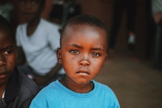 A young child with a serious expression is wearing a blue shirt. The background features other children and a blurred setting that suggests an outdoor or community space.