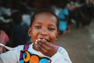 A young child with short hair covers their mouth with one hand, appearing joyful. They are wearing a brightly colored outfit with circular patterns. The background is blurred, showing indistinct figures and a dimly lit environment.
