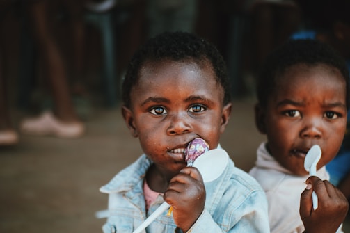 Two young children are looking directly at the camera. One child is holding a spoon and has a lollipop in their mouth, wearing a denim jacket. The second child is in the background, also holding a spoon.