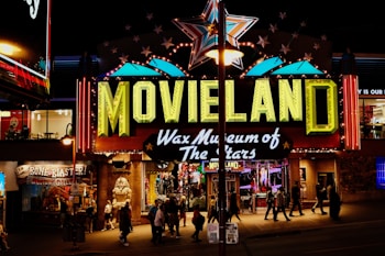 A brightly lit entrance to a wax museum named 'Movieland Wax Museum of The Stars' featuring a large sign with neon lights. A giant star is displayed above the sign and various other illuminated decorations. Several people are walking in front of the entrance, and there are other shops and signs visible around it, adding to the lively night atmosphere.