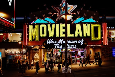 A brightly lit entrance to a wax museum named 'Movieland Wax Museum of The Stars' featuring a large sign with neon lights. A giant star is displayed above the sign and various other illuminated decorations. Several people are walking in front of the entrance, and there are other shops and signs visible around it, adding to the lively night atmosphere.