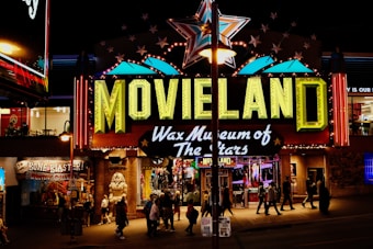 A brightly lit entrance to a wax museum named 'Movieland Wax Museum of The Stars' featuring a large sign with neon lights. A giant star is displayed above the sign and various other illuminated decorations. Several people are walking in front of the entrance, and there are other shops and signs visible around it, adding to the lively night atmosphere.