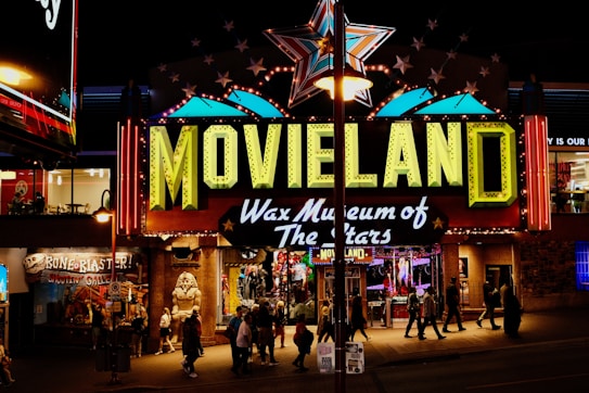 A brightly lit entrance to a wax museum named 'Movieland Wax Museum of The Stars' featuring a large sign with neon lights. A giant star is displayed above the sign and various other illuminated decorations. Several people are walking in front of the entrance, and there are other shops and signs visible around it, adding to the lively night atmosphere.
