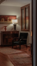 An elegantly furnished room featuring a wooden cabinet with a decorative lamp and an abstract painting on the wall. The setup includes a dark upholstered armchair positioned on a wooden floor with patterned rugs. A window with stained glass detail adds to the sophisticated ambiance.