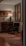 An elegantly furnished room featuring a wooden cabinet with a decorative lamp and an abstract painting on the wall. The setup includes a dark upholstered armchair positioned on a wooden floor with patterned rugs. A window with stained glass detail adds to the sophisticated ambiance.