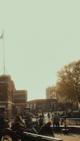 A group of people gathers around a historic brick building with a flagpole. The building is surrounded by a fenced area, and in the background, another structure with columns is visible. A large tree with yellow leaves adds a touch of nature to the urban setting.