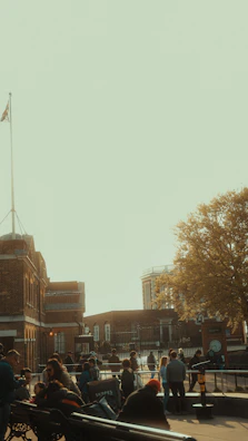 A group of people gathers around a historic brick building with a flagpole. The building is surrounded by a fenced area, and in the background, another structure with columns is visible. A large tree with yellow leaves adds a touch of nature to the urban setting.