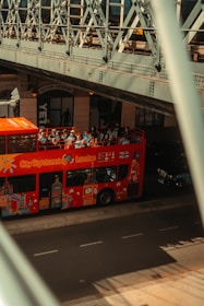 A red City Sightseeing bus with tourists on the top deck passes under a metal bridge. The side of the bus is decorated with iconic London landmarks. The bridge's intricate metal architecture creates shadows on the road. A nearby building has a sign reading 'Ocean Leisure' above the entrance.