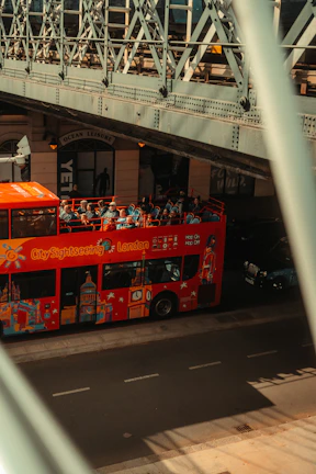 A red City Sightseeing bus with tourists on the top deck passes under a metal bridge. The side of the bus is decorated with iconic London landmarks. The bridge's intricate metal architecture creates shadows on the road. A nearby building has a sign reading 'Ocean Leisure' above the entrance.
