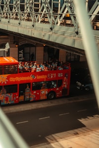 A red City Sightseeing bus with tourists on the top deck passes under a metal bridge. The side of the bus is decorated with iconic London landmarks. The bridge's intricate metal architecture creates shadows on the road. A nearby building has a sign reading 'Ocean Leisure' above the entrance.