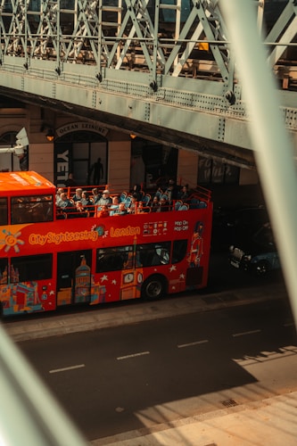 A red City Sightseeing bus with tourists on the top deck passes under a metal bridge. The side of the bus is decorated with iconic London landmarks. The bridge's intricate metal architecture creates shadows on the road. A nearby building has a sign reading 'Ocean Leisure' above the entrance.
