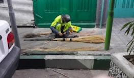 A person is sitting on the ground, assembling several straw brooms. They appear to be working diligently, with multiple broom heads and handles laid out on the ground around them. Nearby, there is a green gate, a white vehicle, and some plants.