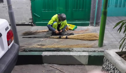 A person is sitting on the ground, assembling several straw brooms. They appear to be working diligently, with multiple broom heads and handles laid out on the ground around them. Nearby, there is a green gate, a white vehicle, and some plants.