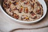 A baked dessert placed in an oval ceramic dish is topped with slices of fresh figs and sprinkled with sesame seeds. The dish rests on a fabric with a neutral color, creating a rustic and warm presentation.