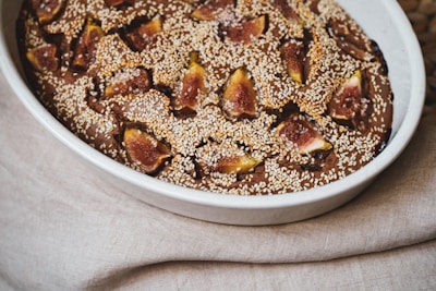 A baked dessert placed in an oval ceramic dish is topped with slices of fresh figs and sprinkled with sesame seeds. The dish rests on a fabric with a neutral color, creating a rustic and warm presentation.