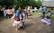 A group of people is engaged in a lively outdoor dance, dressed in traditional clothing, with trees and a wooden house in the background. The scene is vibrant and full of movement.