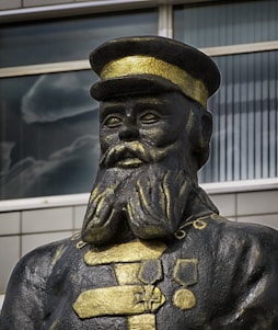 A statue of a man wearing a military uniform with a hat. The uniform is decorated with medals and has a prominent texture, colored in shades of black and gold. The background shows a modern building with large windows.