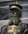 A statue of a man wearing a military uniform with a hat. The uniform is decorated with medals and has a prominent texture, colored in shades of black and gold. The background shows a modern building with large windows.