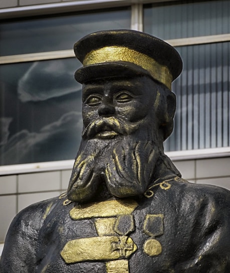 A statue of a man wearing a military uniform with a hat. The uniform is decorated with medals and has a prominent texture, colored in shades of black and gold. The background shows a modern building with large windows.