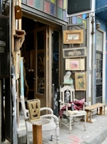 An eclectic storefront display with vintage wooden chairs and framed pictures. The chairs are placed on either side of the entrance, one holding a folded red cloth. Various artworks and items are hung on the walls around the door, with a mix of portrait and abstract styles. The building features a colorful stained-glass panel above the doorway, and the surrounding walls are painted in different shades, creating a vibrant, artistic atmosphere.