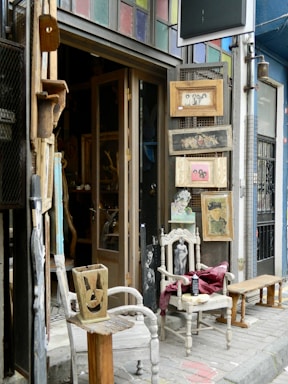 An eclectic storefront display with vintage wooden chairs and framed pictures. The chairs are placed on either side of the entrance, one holding a folded red cloth. Various artworks and items are hung on the walls around the door, with a mix of portrait and abstract styles. The building features a colorful stained-glass panel above the doorway, and the surrounding walls are painted in different shades, creating a vibrant, artistic atmosphere.