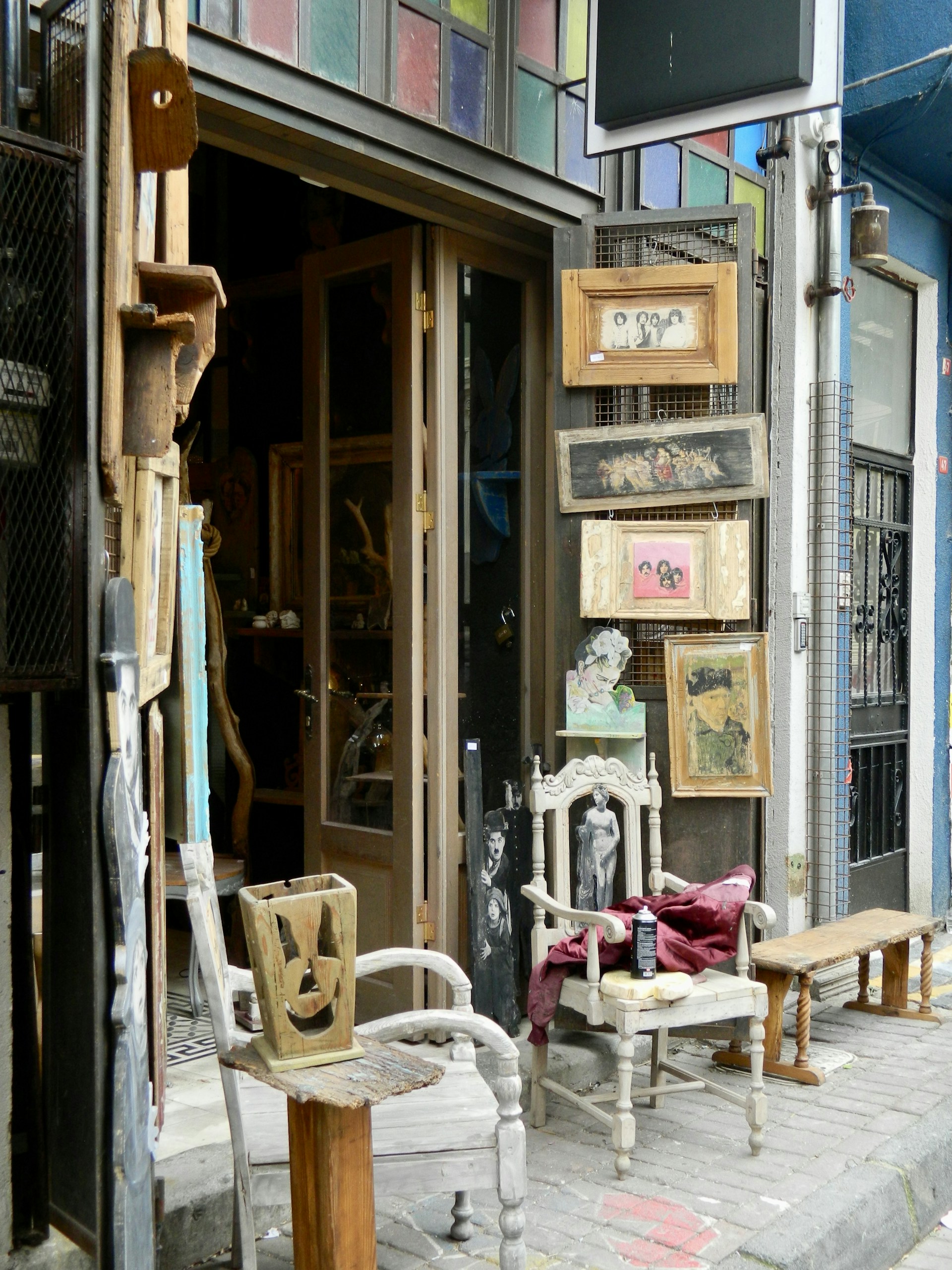 An eclectic storefront display with vintage wooden chairs and framed pictures. The chairs are placed on either side of the entrance, one holding a folded red cloth. Various artworks and items are hung on the walls around the door, with a mix of portrait and abstract styles. The building features a colorful stained-glass panel above the doorway, and the surrounding walls are painted in different shades, creating a vibrant, artistic atmosphere.