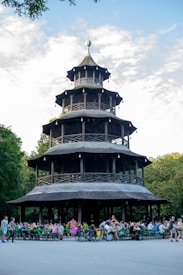 A large, multi-tiered wooden pagoda surrounded by trees serves as the focal point. Below, numerous people are seated at green picnic tables, enjoying an outdoor social gathering. The scene is set in a lush park with a partially cloudy sky overhead.