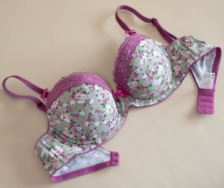 A floral-patterned bra with pink lace detailing and purple straps, laid flat on a beige surface. The cups and band feature a light green fabric adorned with small pink and white flowers.