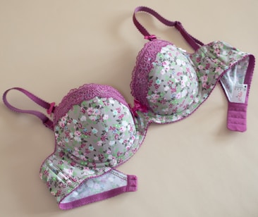 A floral-patterned bra with pink lace detailing and purple straps, laid flat on a beige surface. The cups and band feature a light green fabric adorned with small pink and white flowers.