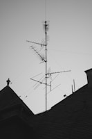 A rooftop with a large metal antenna structure, featuring multiple elements and wires extending outward. The silhouette of the antenna contrasts against a clear sky, while the roof's textured shingles create an interesting pattern.