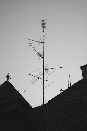 A rooftop with a large metal antenna structure, featuring multiple elements and wires extending outward. The silhouette of the antenna contrasts against a clear sky, while the roof's textured shingles create an interesting pattern.