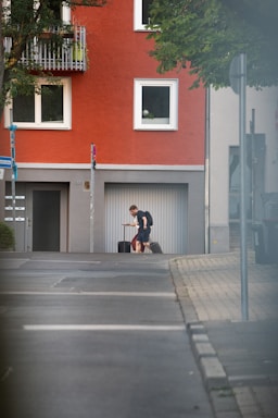 A person with a backpack is walking down a street while looking at a mobile device. There is a suitcase nearby. The backdrop features a vibrant orange building with multiple windows and a balcony with greenery. The foreground shows part of a road with sidewalks and a traffic sign.