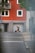 A person with a backpack is walking down a street while looking at a mobile device. There is a suitcase nearby. The backdrop features a vibrant orange building with multiple windows and a balcony with greenery. The foreground shows part of a road with sidewalks and a traffic sign.