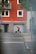 A person with a backpack is walking down a street while looking at a mobile device. There is a suitcase nearby. The backdrop features a vibrant orange building with multiple windows and a balcony with greenery. The foreground shows part of a road with sidewalks and a traffic sign.