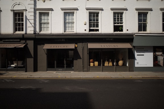 The image captures a storefront with large windows displaying various decorative items like vases and small furniture pieces. The facade has a classic architectural design with white upper walls and large, vertical windows situated above the black-framed shop displays. The street in front of the store is empty, and a billboard on the side advertises 'London Design Week'.