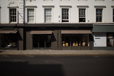The image captures a storefront with large windows displaying various decorative items like vases and small furniture pieces. The facade has a classic architectural design with white upper walls and large, vertical windows situated above the black-framed shop displays. The street in front of the store is empty, and a billboard on the side advertises 'London Design Week'.