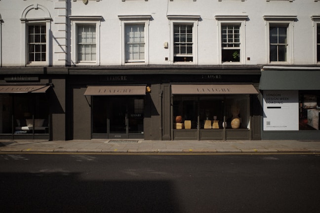 The image captures a storefront with large windows displaying various decorative items like vases and small furniture pieces. The facade has a classic architectural design with white upper walls and large, vertical windows situated above the black-framed shop displays. The street in front of the store is empty, and a billboard on the side advertises 'London Design Week'.