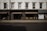 The image captures a storefront with large windows displaying various decorative items like vases and small furniture pieces. The facade has a classic architectural design with white upper walls and large, vertical windows situated above the black-framed shop displays. The street in front of the store is empty, and a billboard on the side advertises 'London Design Week'.
